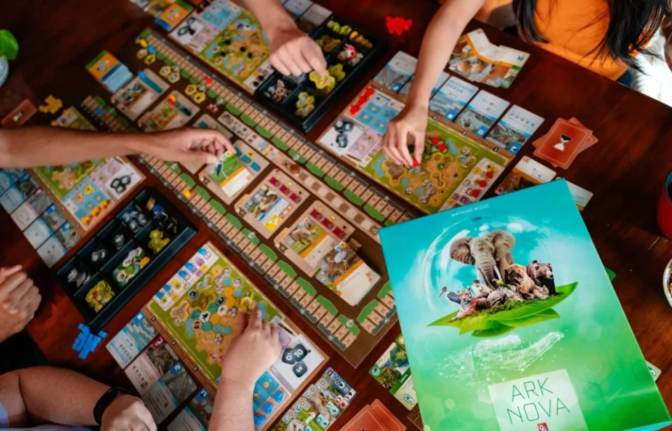 Top-down Ark Nova table setup with central board, player zoo maps, cards, and component trays