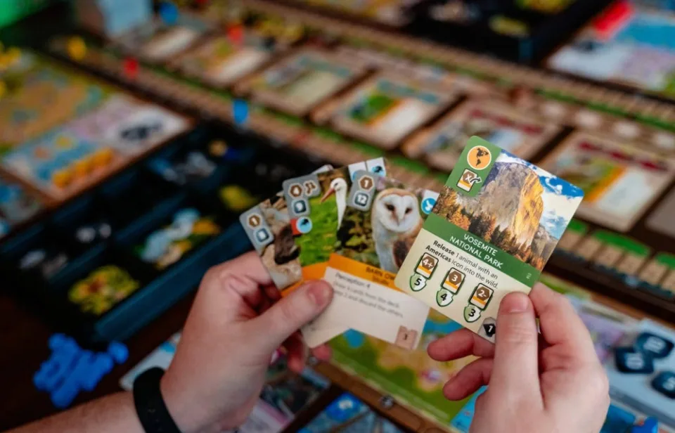 Ark Nova animal and conservation cards held above a player zoo map
